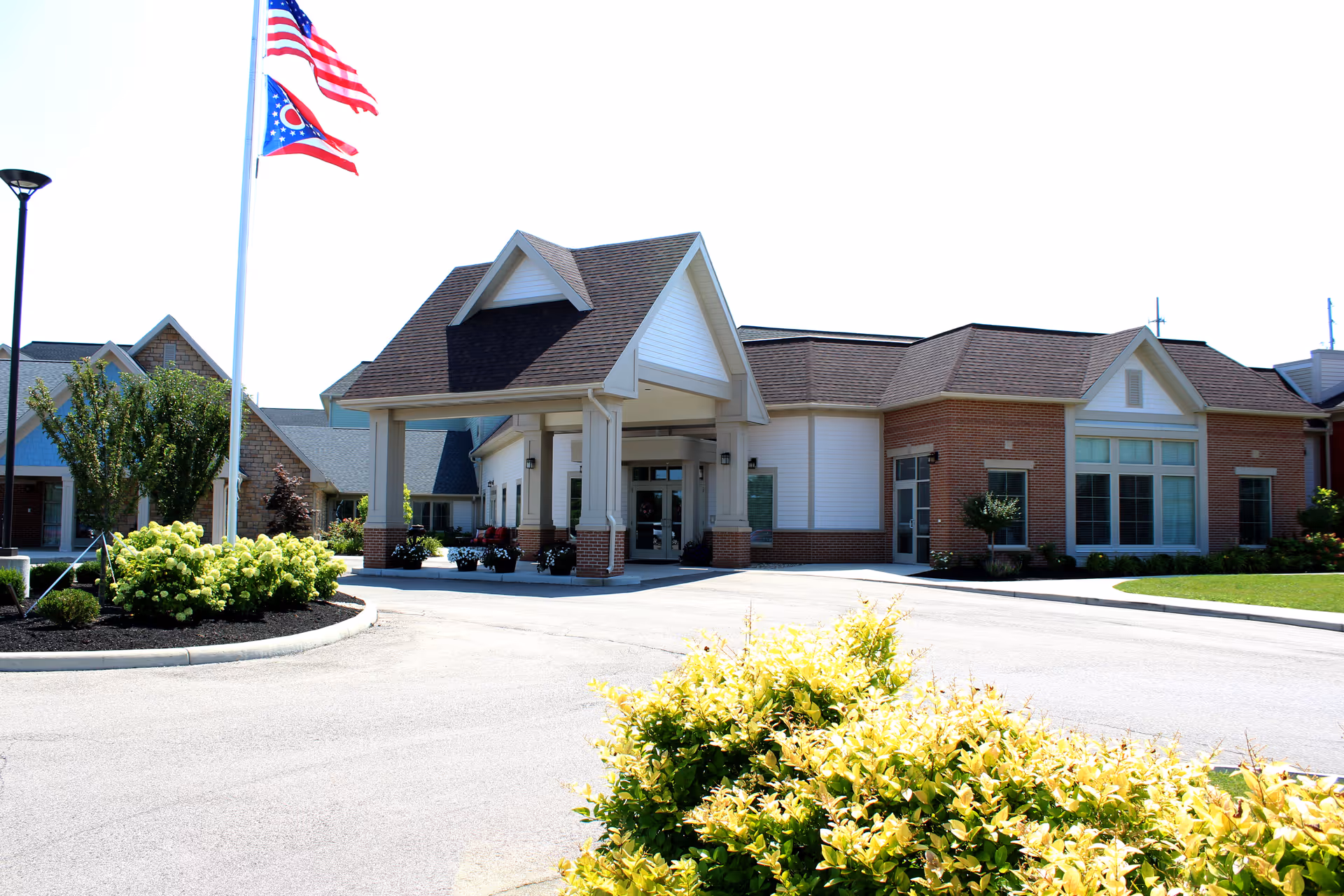Front entrance of the Village at the Greene senior living building with a covered porte-cochère, flagpoles, and landscaped beds.