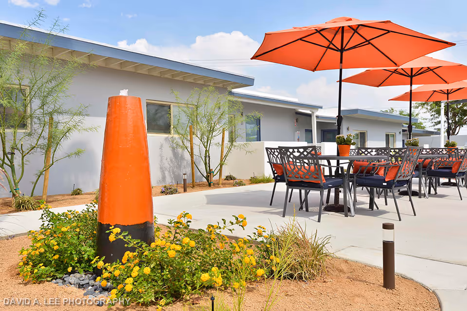 Outdoor patio with orange umbrellas, metal tables and chairs, landscaping, and a single-story building in the background.