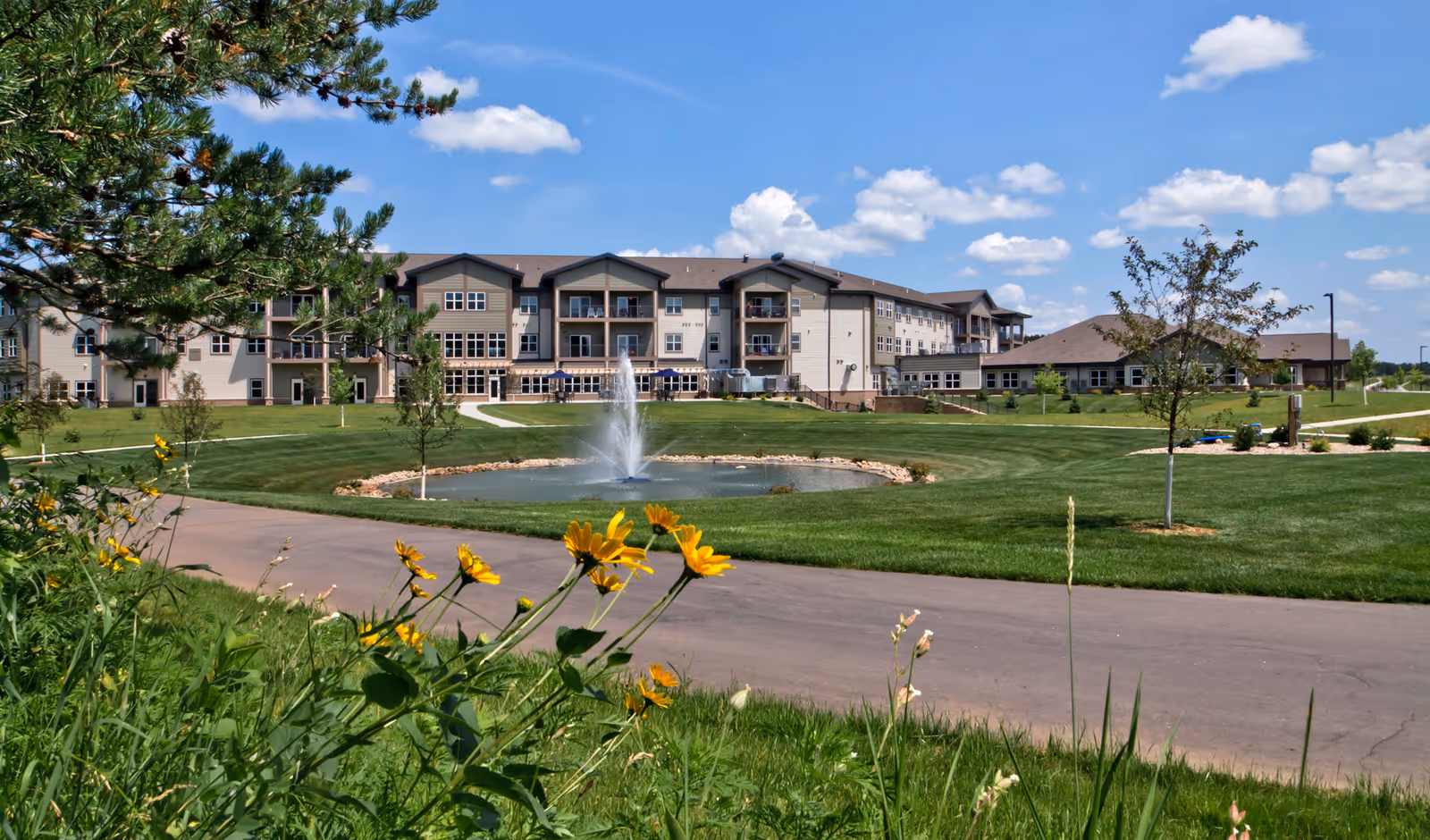 A large senior living facility building with multiple floors and balconies, surrounded by a well-maintained lawn and a paved walking path. In the foreground, there are yellow flowers and green grass, with a small pond featuring a water fountain in the center. The sky is blue with scattered white clouds.