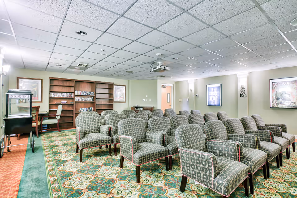 A small theater room with several rows of patterned armchairs facing a projector mounted on the ceiling. The room has a patterned carpet, bookshelves with DVDs, framed movie posters on the walls, and a popcorn machine in the corner.