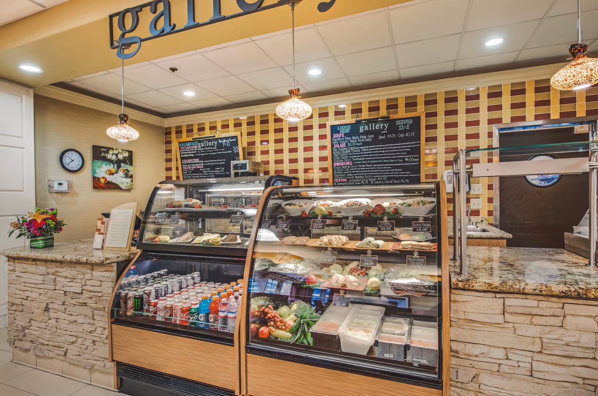 Interior view of a small café or deli counter with glass display cases filled with various food items and beverages. Behind the counter, there are two menu boards listing breakfast and lunch options. The counter has a stone facade with a granite countertop, and there are hanging pendant lights above. A clock and a framed picture are visible on the wall to the left.
