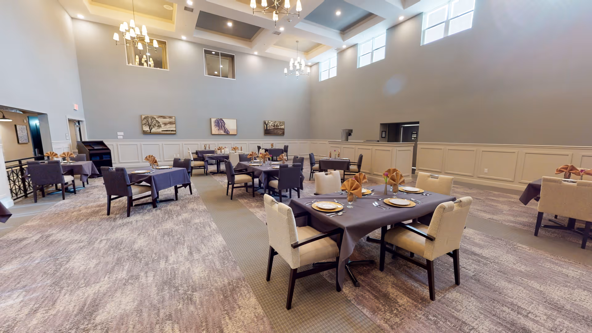 Spacious dining room with multiple tables covered in dark tablecloths, each set with plates, utensils, and folded napkins. The room has high ceilings with chandeliers, large windows near the ceiling allowing natural light, and neutral-colored walls with decorative paneling. Artwork is displayed on the walls, and the carpet has a patterned design.