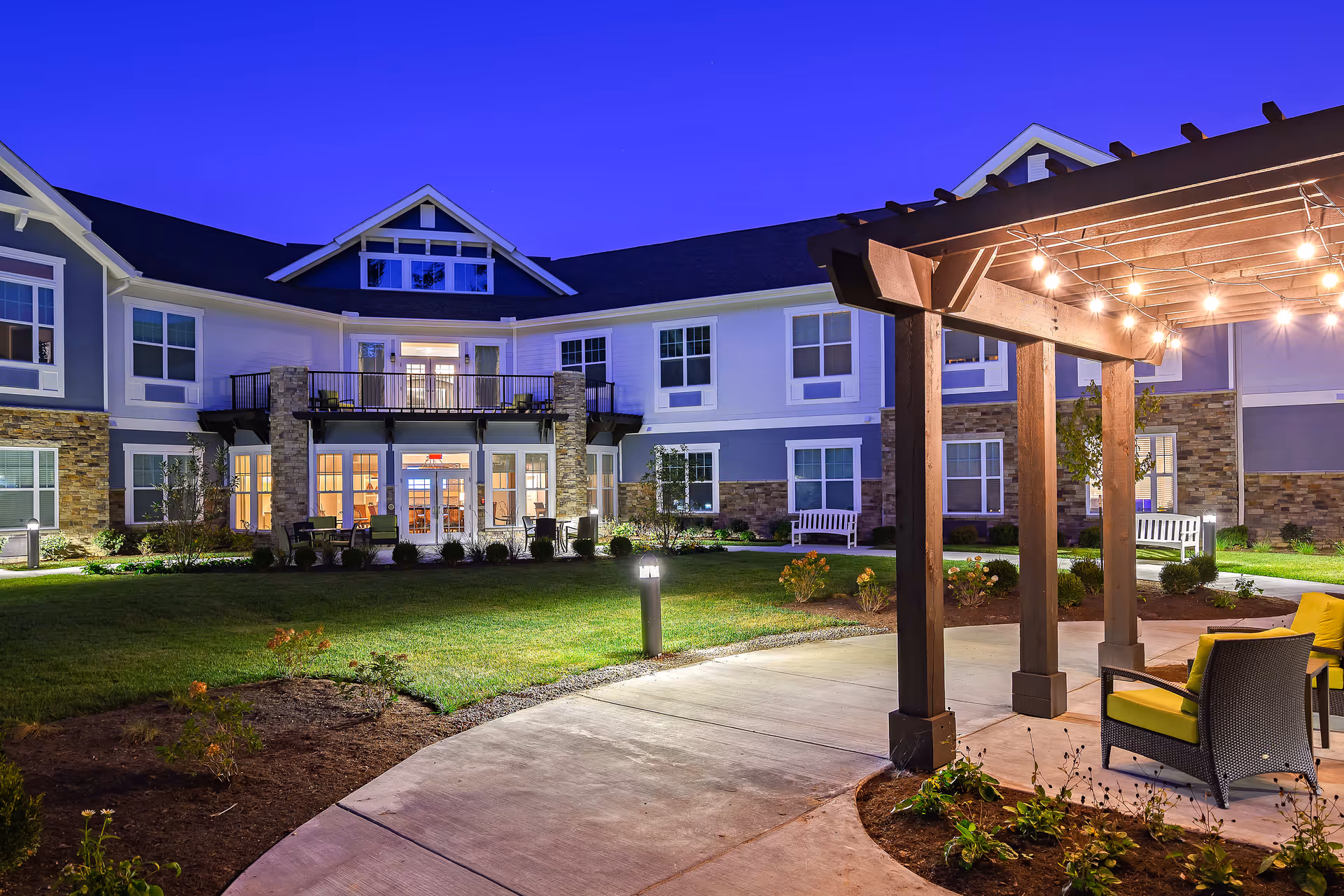 Evening view of The Ashton at Anderson senior living facility showing a well-lit outdoor patio area with a wooden pergola adorned with string lights, comfortable seating with yellow cushions, a curved concrete walkway, landscaped garden beds, and the two-story building with large windows and balconies in the background.