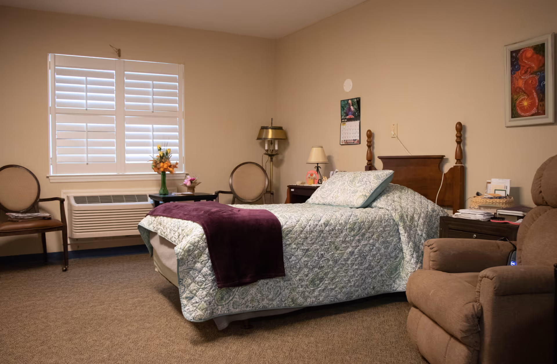A cozy bedroom with a single bed covered in a patterned quilt and a purple throw blanket. The room has beige walls, a window with white shutters, two chairs, a small table with a vase of flowers, a floor lamp, a nightstand with a table lamp, and a comfortable recliner chair. There is a calendar and framed artwork on the walls.