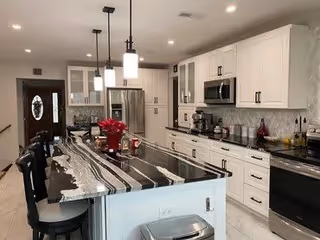Modern kitchen interior with white cabinetry, stainless steel appliances, and a large island with a black and white marble countertop. Three black bar stools are positioned at the island, and pendant lights hang above. The kitchen has a tiled backsplash and a door with an oval glass window in the background.