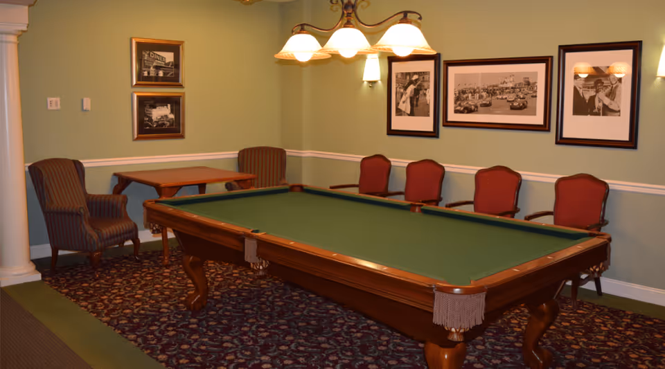 A room with a green felt pool table in the center, surrounded by several red cushioned chairs along the wall. The walls are painted light green and decorated with framed black and white photographs. There is a hanging light fixture above the pool table and a patterned carpet on the floor.