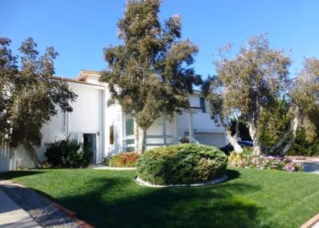 Exterior view of a white two-story residential building surrounded by green grass, bushes, and several trees under a clear blue sky.