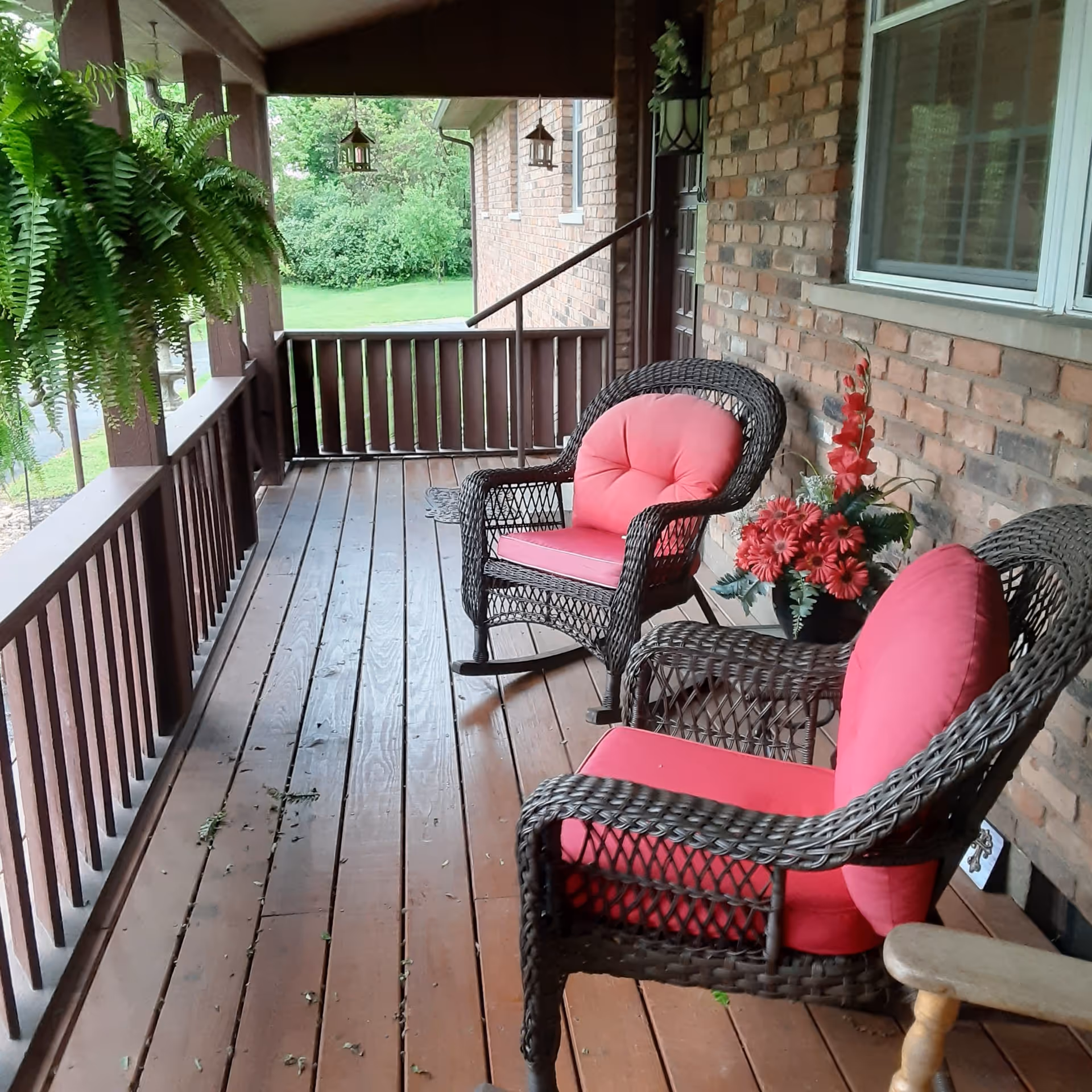 A covered wooden porch with two wicker chairs featuring pink cushions, a small wicker table with a flower arrangement, hanging green fern plants, and a brick wall with a window and door.