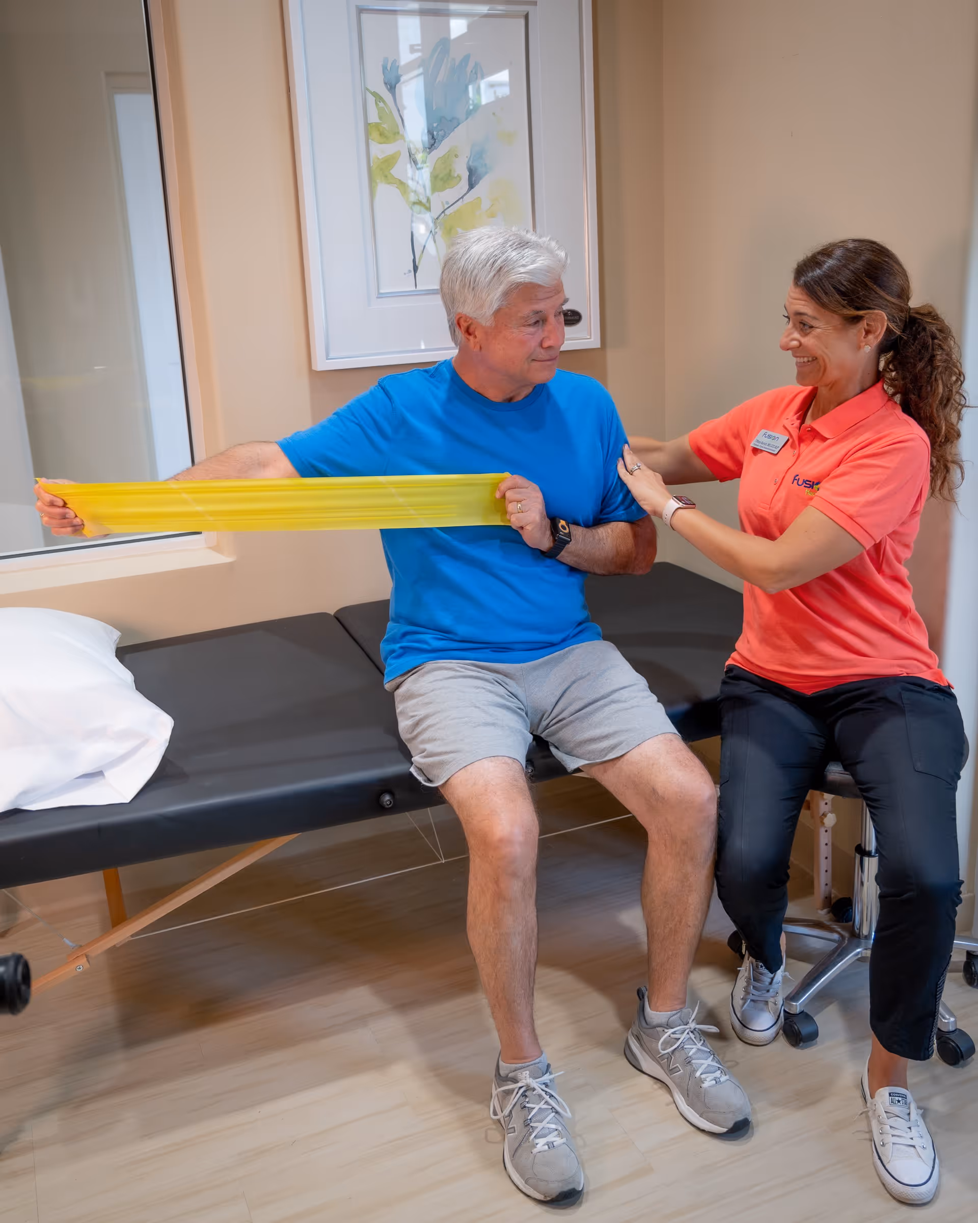 An elderly man in a blue shirt and gray shorts is seated on a black examination table, stretching a yellow resistance band with the assistance of a smiling female therapist wearing a coral polo shirt and black pants in a well-lit room with beige walls and a framed abstract painting.