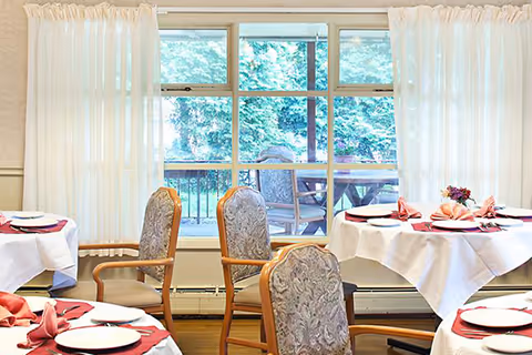 Dining area with round tables covered in white tablecloths and set with plates, napkins, and silverware. Upholstered chairs surround the tables. Large windows with sheer white curtains reveal an outdoor patio with additional seating and greenery.