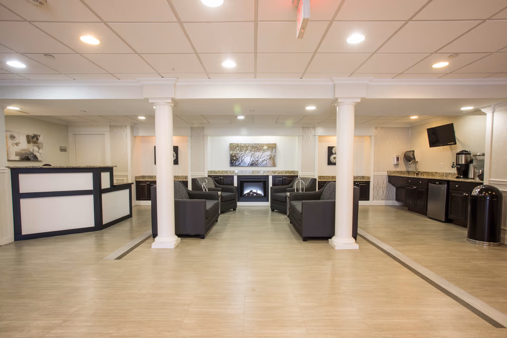A spacious and well-lit common area in a senior living facility featuring a seating area with four black armchairs arranged around a modern electric fireplace. The room has white columns, a reception desk on the left, and a kitchenette area with a coffee machine, sink, and small refrigerator on the right. The ceiling has recessed lighting, and there are decorative artworks on the walls.