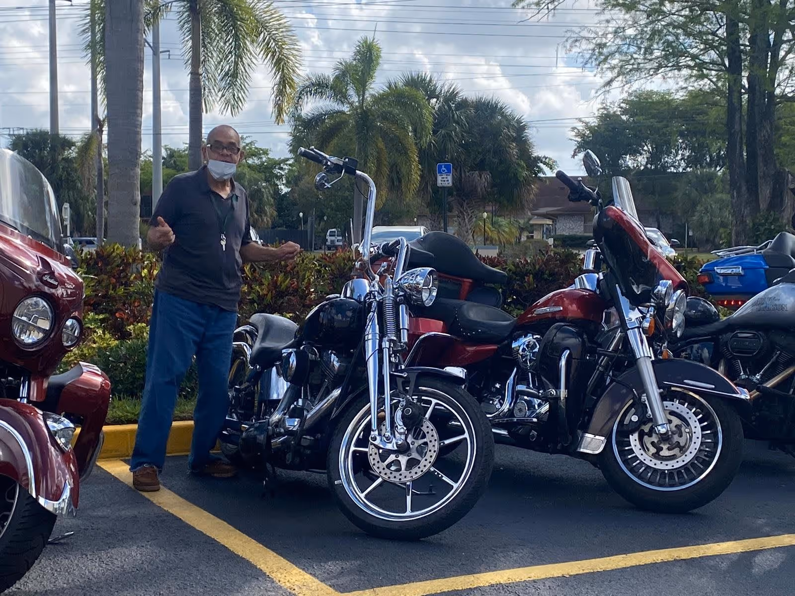 An elderly man wearing a mask and glasses stands in a parking lot next to several parked motorcycles. Palm trees and bushes are visible in the background under a partly cloudy sky.