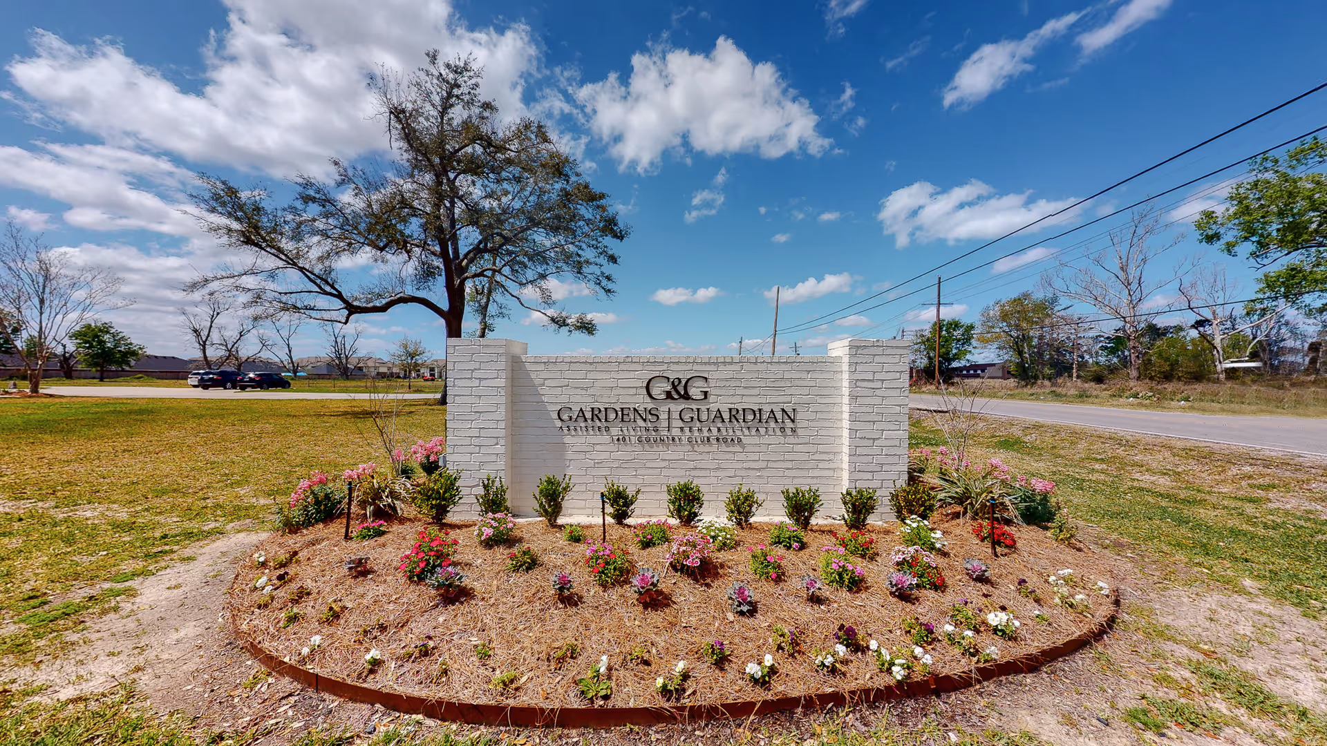 White brick entrance sign for The Gardens & Guardian surrounded by a circular flowerbed on a grassy lot under a blue sky.