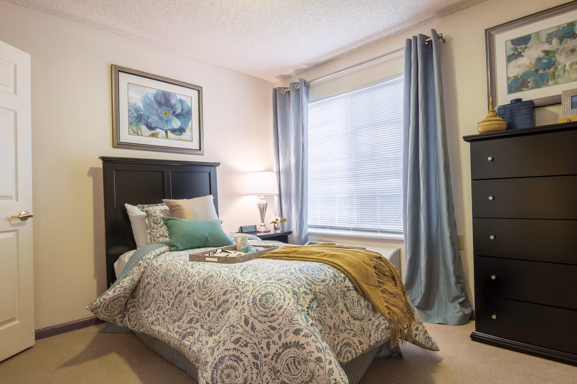 Well-lit bedroom with a patterned bed, teal and yellow accents, a nightstand with lamp, and a tall dresser by the window.