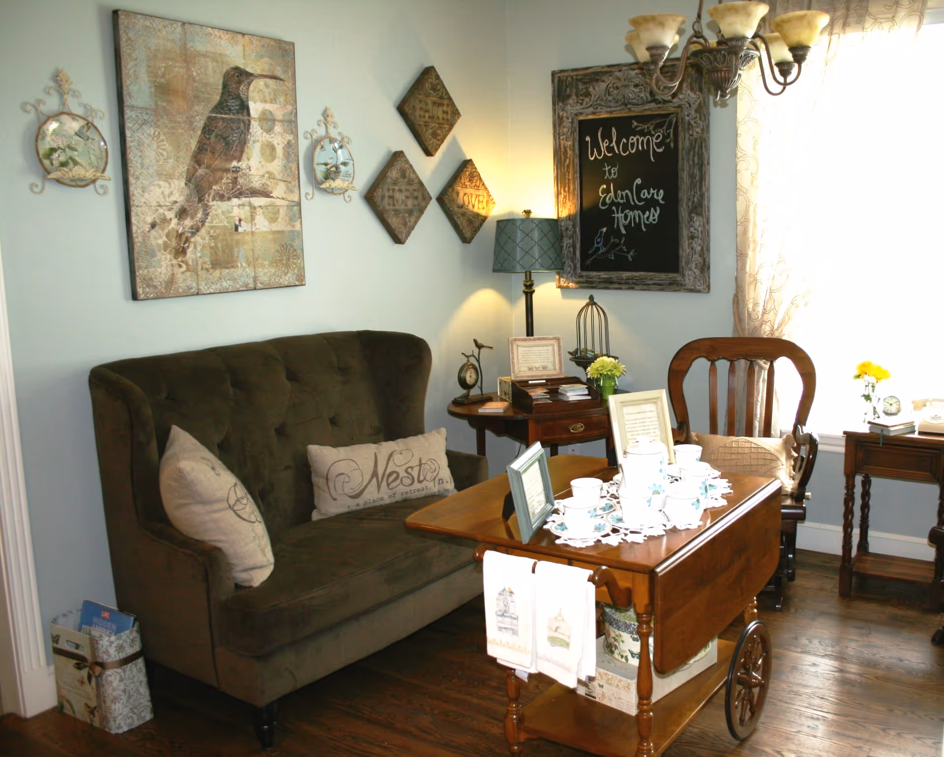 A cozy living room area with a dark green tufted loveseat adorned with decorative pillows, a wooden tea cart set with a white tea set, and a wooden chair. The walls are decorated with framed artwork including a large bird painting, diamond-shaped plaques with words like 'Faith', 'Hope', and 'Love', and a chalkboard sign that reads 'Welcome to Eden Care Homes'. A table lamp and small side tables with flowers and framed pictures complete the scene.