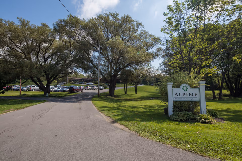 Entrance road leading to Alpine Rehabilitation & Nursing Center with a sign on the right side surrounded by greenery and trees. Several cars are parked in a parking lot visible in the background under a partly cloudy sky.
