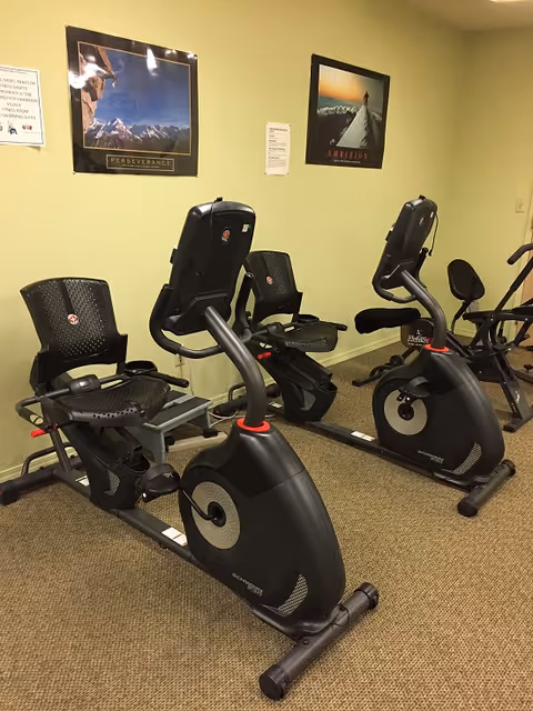 Indoor exercise room with two stationary recumbent bikes and other fitness equipment against a beige wall with motivational posters.