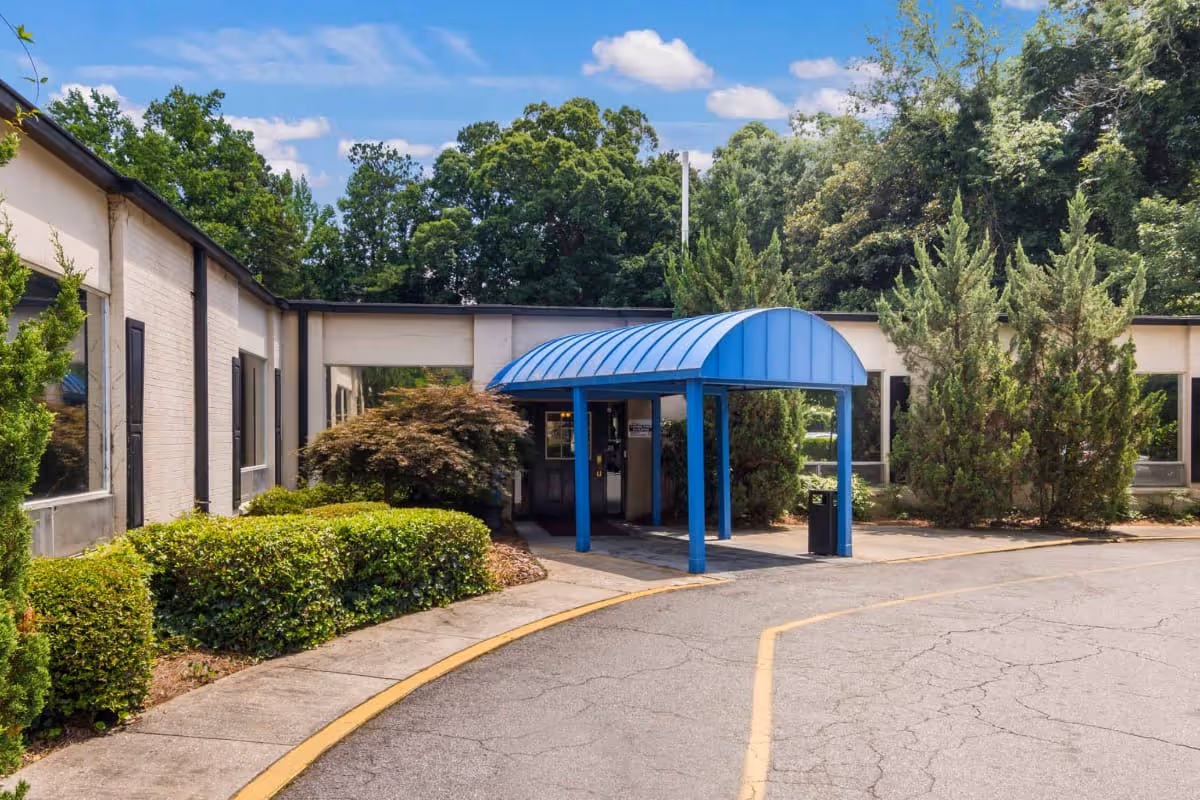 Exterior view of Harborview Decatur facility entrance with a blue covered walkway, surrounded by green bushes and trees under a partly cloudy sky.
