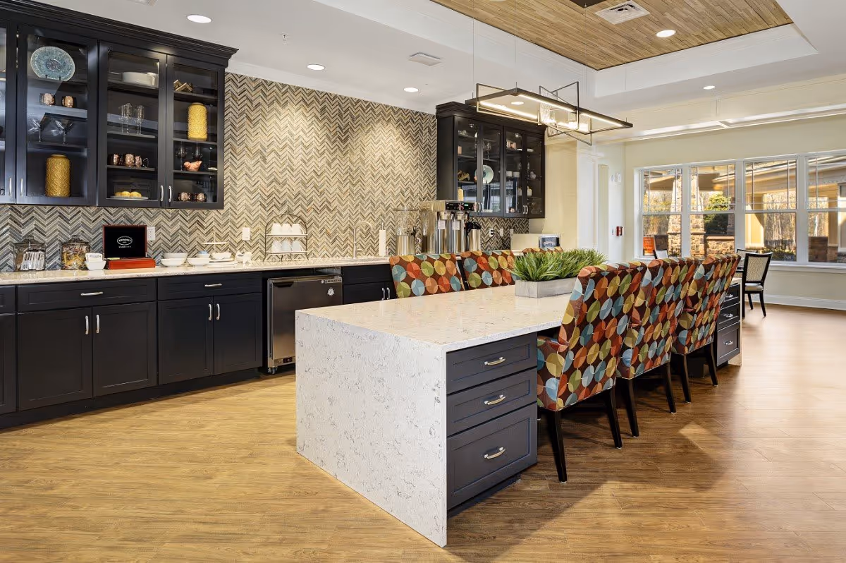 A modern kitchen area with a large white marble island countertop surrounded by colorful patterned chairs. The back wall features dark cabinets with glass doors displaying dishes and decorative items, and a chevron-patterned backsplash. The floor is wooden, and large windows let in natural light.