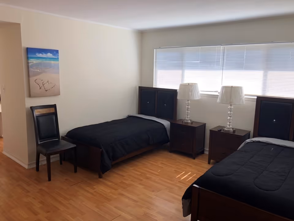 A bedroom with two single beds featuring dark wooden frames and black bedding. Between the beds are two matching dark wooden nightstands, each with a clear glass lamp with a white lampshade. A large window with white blinds is behind the beds, letting in natural light. There is a black chair against the left wall and a beach-themed painting hanging above it. The floor is light wood.