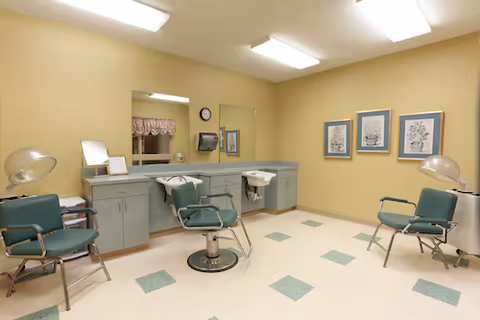 Interior of a hair salon area with three green salon chairs, two hair washing sinks, large mirrors, and cabinets under the mirrors. The walls are painted beige and decorated with three framed pictures. The floor has a light-colored tile with green square accents.