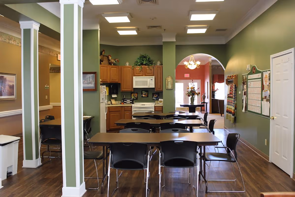 Interior view of a senior living facility dining area with green walls, wooden flooring, and several tables and black chairs arranged in rows. In the background, there is a kitchen area with wooden cabinets, a white refrigerator, microwave, and stove. The room is well-lit with ceiling lights, and there is an archway leading to another room with a chandelier and floral arrangements.