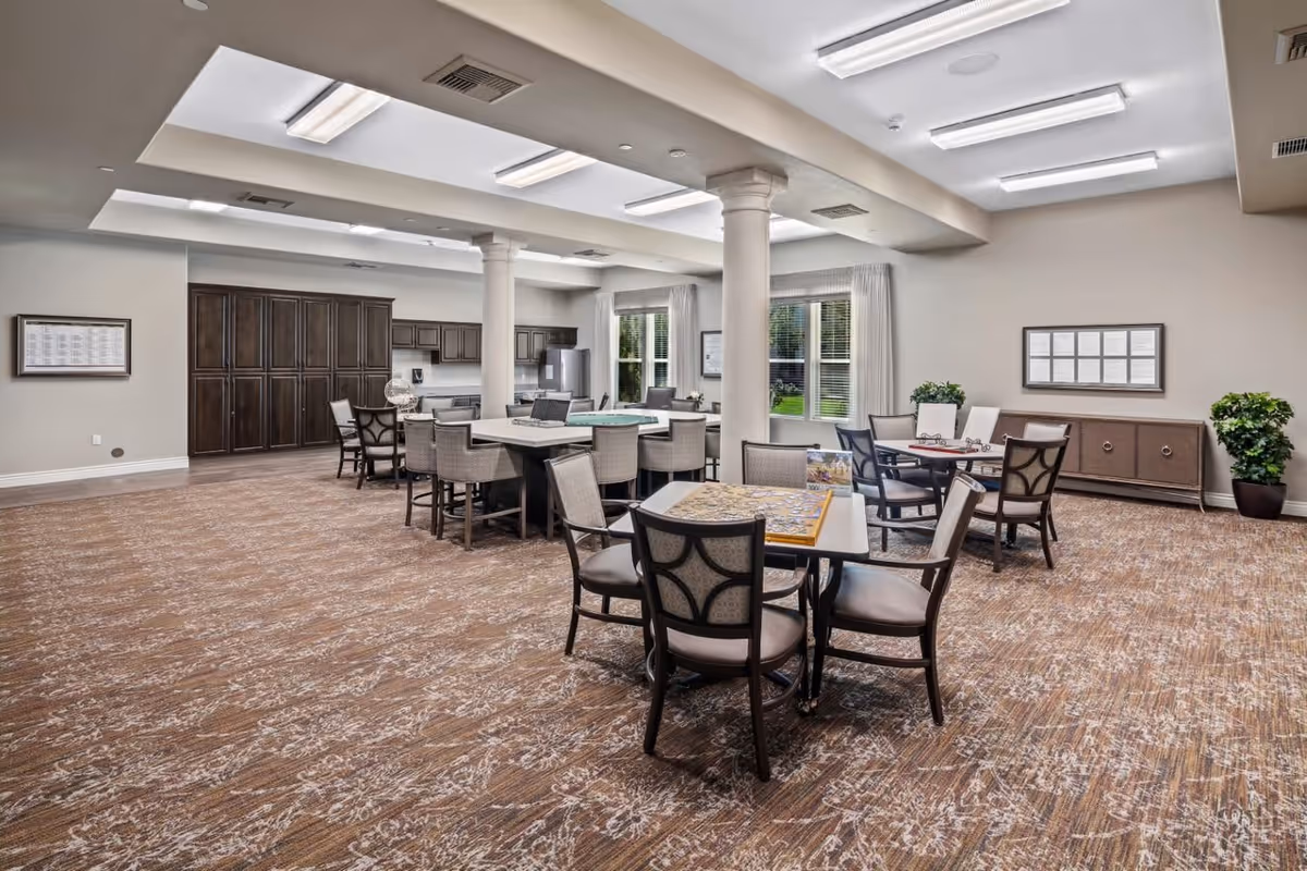 A spacious, well-lit common area with multiple tables and chairs arranged for group activities or dining. The room features large windows with white curtains, two white columns, a patterned carpet, and a kitchen area with dark wood cabinets and stainless steel appliances in the background. There are framed pictures on the walls and potted plants adding greenery to the space.