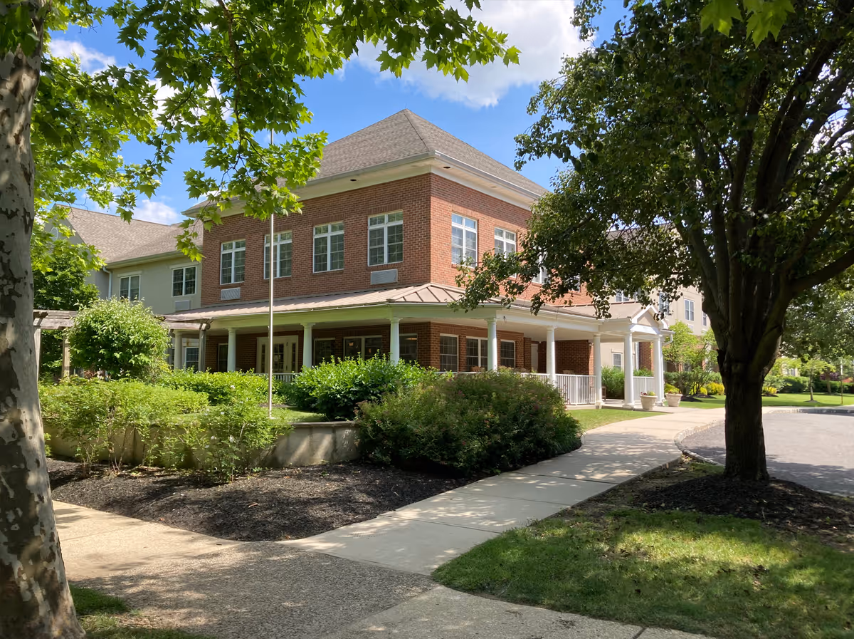 Two-story red brick senior living building with a covered porch, landscaped shrubs, trees, and a sidewalk leading to the entrance.