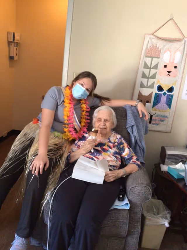 An elderly woman sitting in a gray armchair holding an ice cream cone and a piece of paper, smiling. A younger woman wearing a face mask, a colorful lei, and a grass skirt is leaning on the armchair next to her. The room has light-colored walls with a decorative wall hanging featuring animal designs, a small table with tissues, and a trash bin nearby.