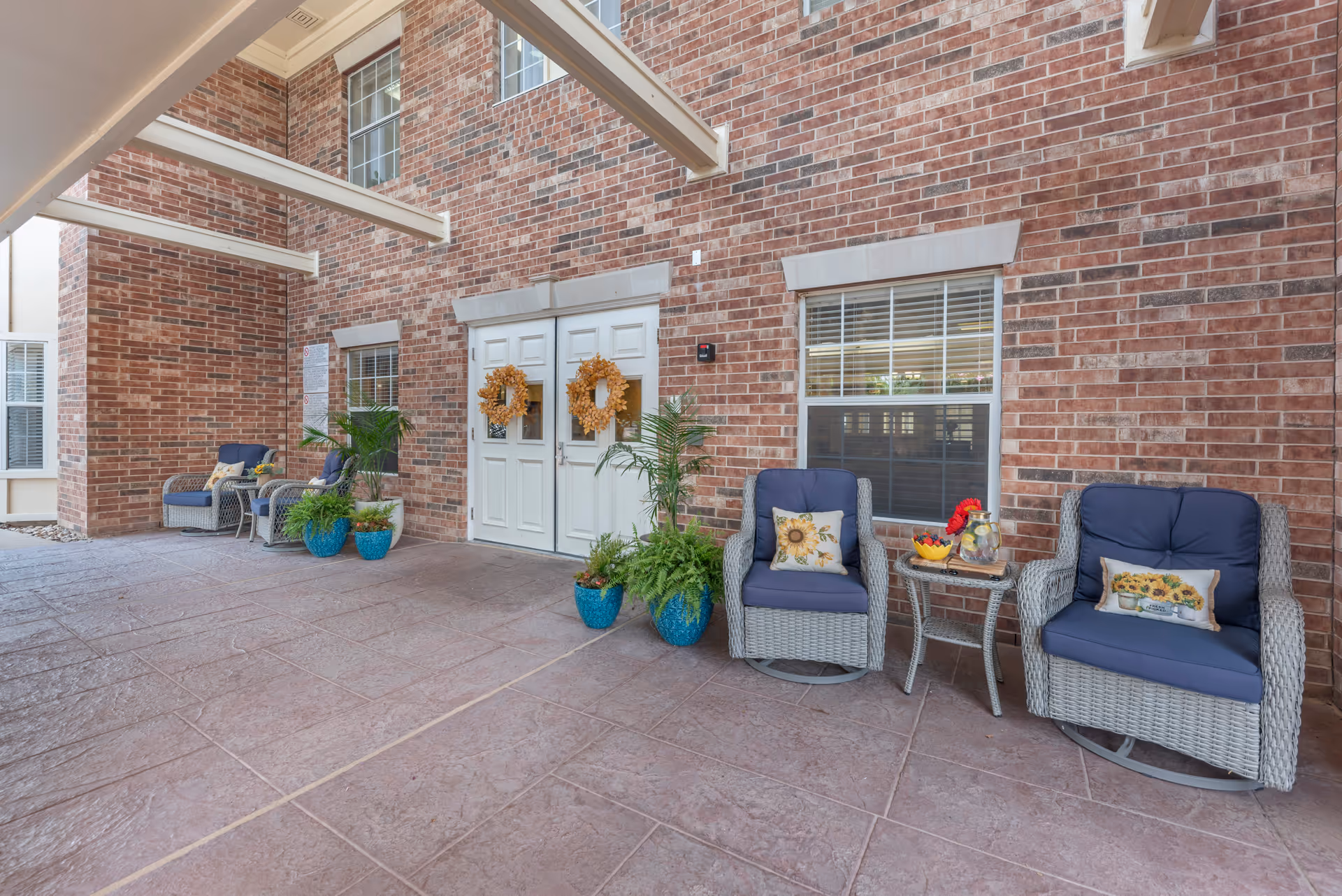 Covered outdoor seating area with four cushioned wicker chairs, two small tables, potted plants, and double doors decorated with wreaths on a brick wall.