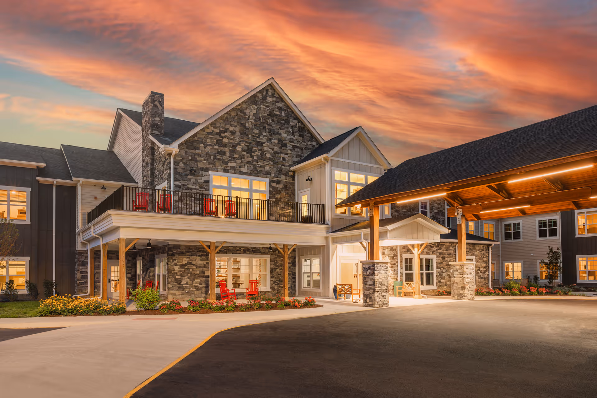 Front exterior of a senior living facility with stone facade, covered porte-cochere, balconies and lit windows at sunset.