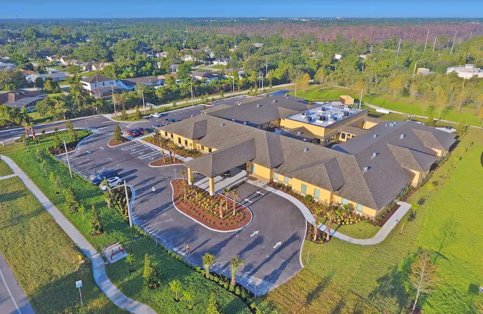 Aerial view of a single-story assisted living and memory care facility with a large parking lot, landscaped entrance, and surrounding greenery. The building has a tan exterior with a dark roof and is situated in a suburban area with trees and houses nearby.