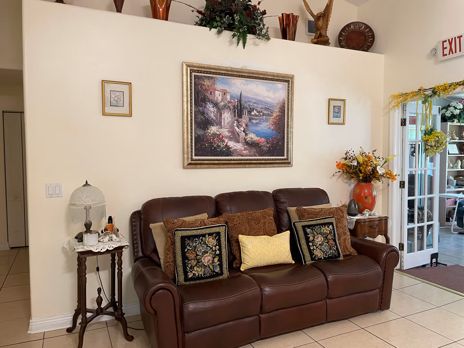 A cozy living room area with a brown leather sofa adorned with multiple decorative pillows. Above the sofa hangs a large framed painting depicting a scenic landscape with flowers, a pathway, and a body of water. On either side of the painting are two smaller framed pictures. To the left of the sofa is a small wooden side table with a vintage-style lamp and some small decorative items. To the right is another small wooden side table with a large red vase filled with artificial flowers. The room has tiled flooring, cream-colored walls, and a glass door decorated with yellow flowers leading to another room. An exit sign is visible above the door.
