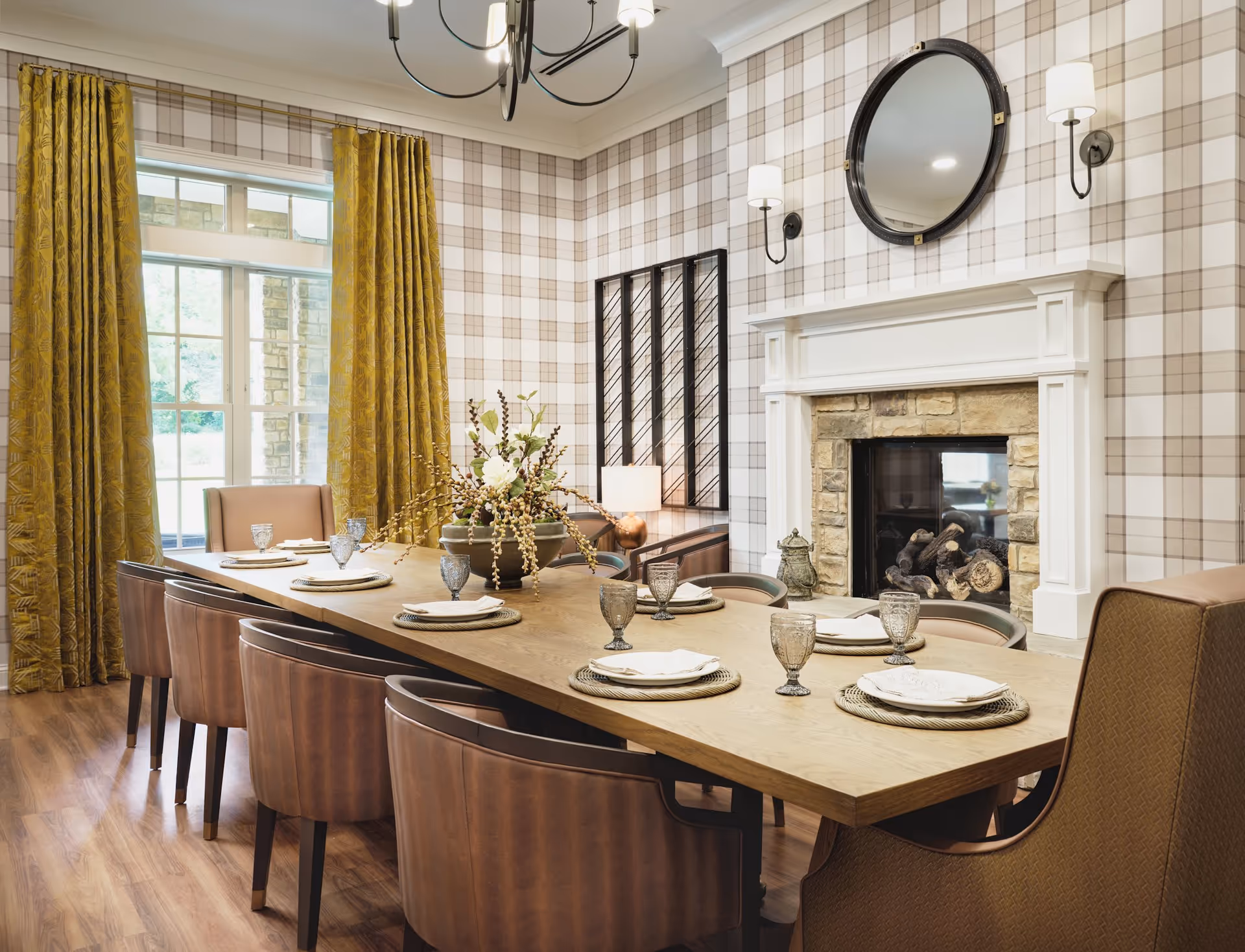 A dining room with a long wooden table set with plates, napkins, and glasses. The room features brown leather chairs, a stone fireplace with a white mantel, a round mirror above the fireplace, plaid wallpaper, mustard yellow curtains, and a chandelier hanging from the ceiling.