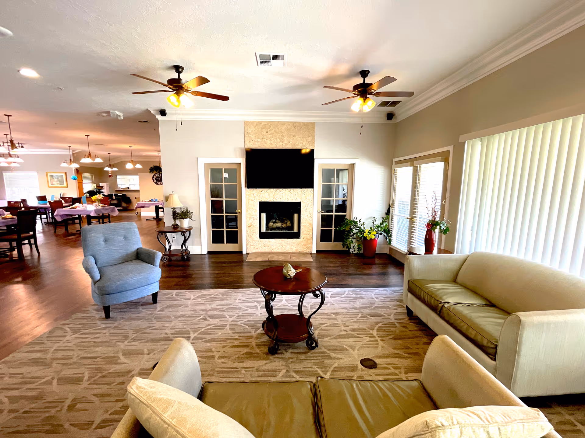 Bright communal living room with sofas, an armchair, a coffee table, fireplace and TV, with a dining area visible in the background.