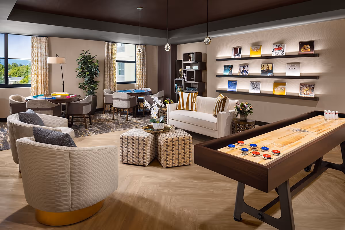 A cozy senior living common area with a shuffleboard table in the foreground, two beige armchairs with gray pillows, a white sofa with striped pillows, two patterned ottomans, and two round tables with chairs near large windows. The walls feature shelves displaying vinyl records and a modern bookshelf with decorative items. The room has warm lighting and a mix of wood and carpet flooring.