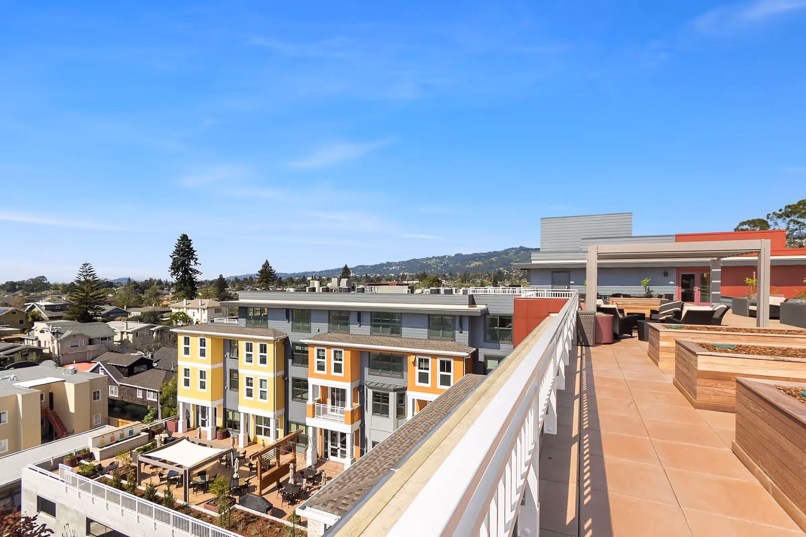 View from a rooftop terrace overlooking a modern multi-story residential building with colorful facades in yellow, orange, and gray. The terrace features wooden planters, outdoor seating, and a pergola under a clear blue sky with distant hills in the background.