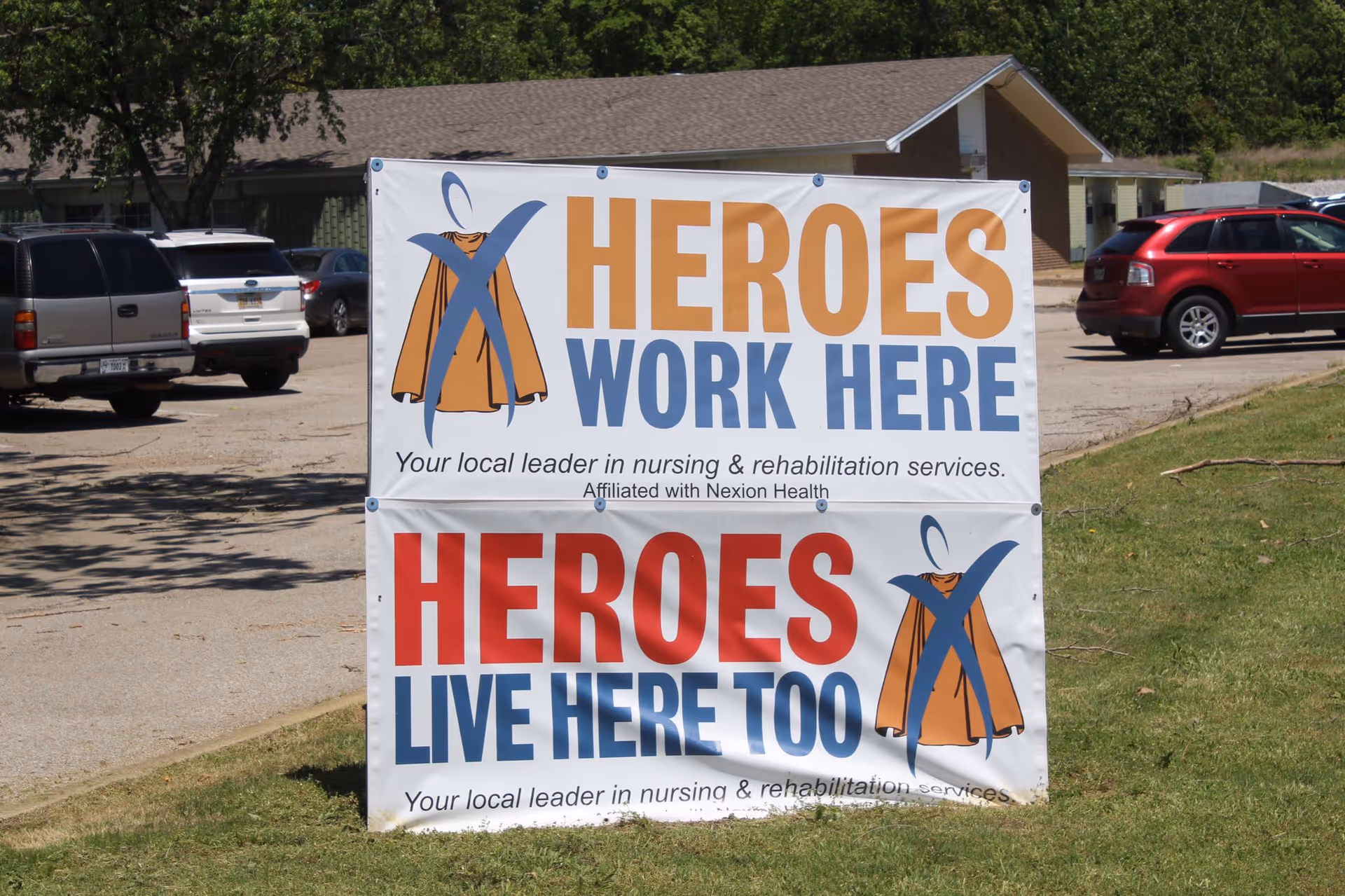 Outdoor sign on a lawn reading 'HEROES WORK HERE' and 'HEROES LIVE HERE TOO' with cape graphics, with parked cars and a building behind it.