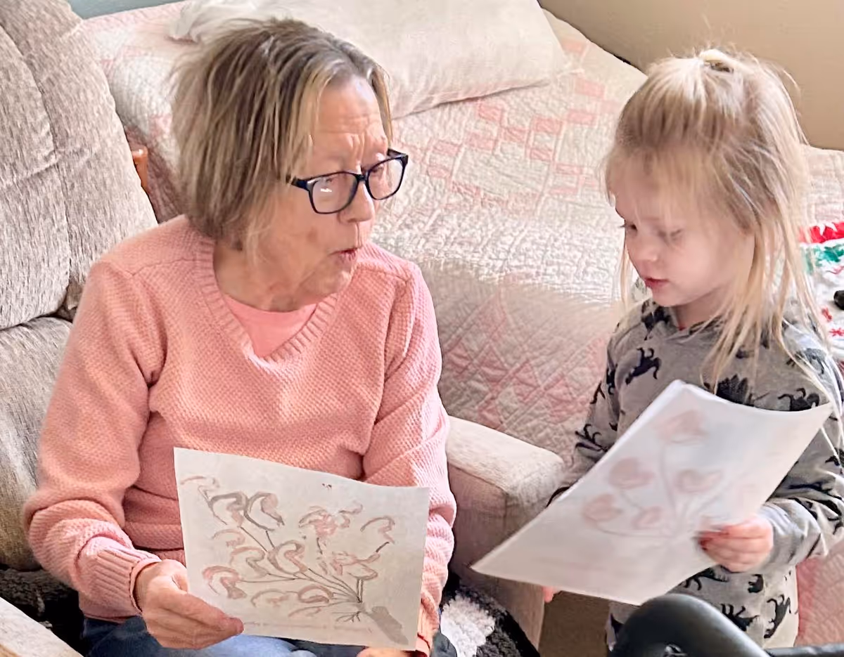 An elderly woman and a young girl sit indoors holding drawings with a quilted bed visible behind them.