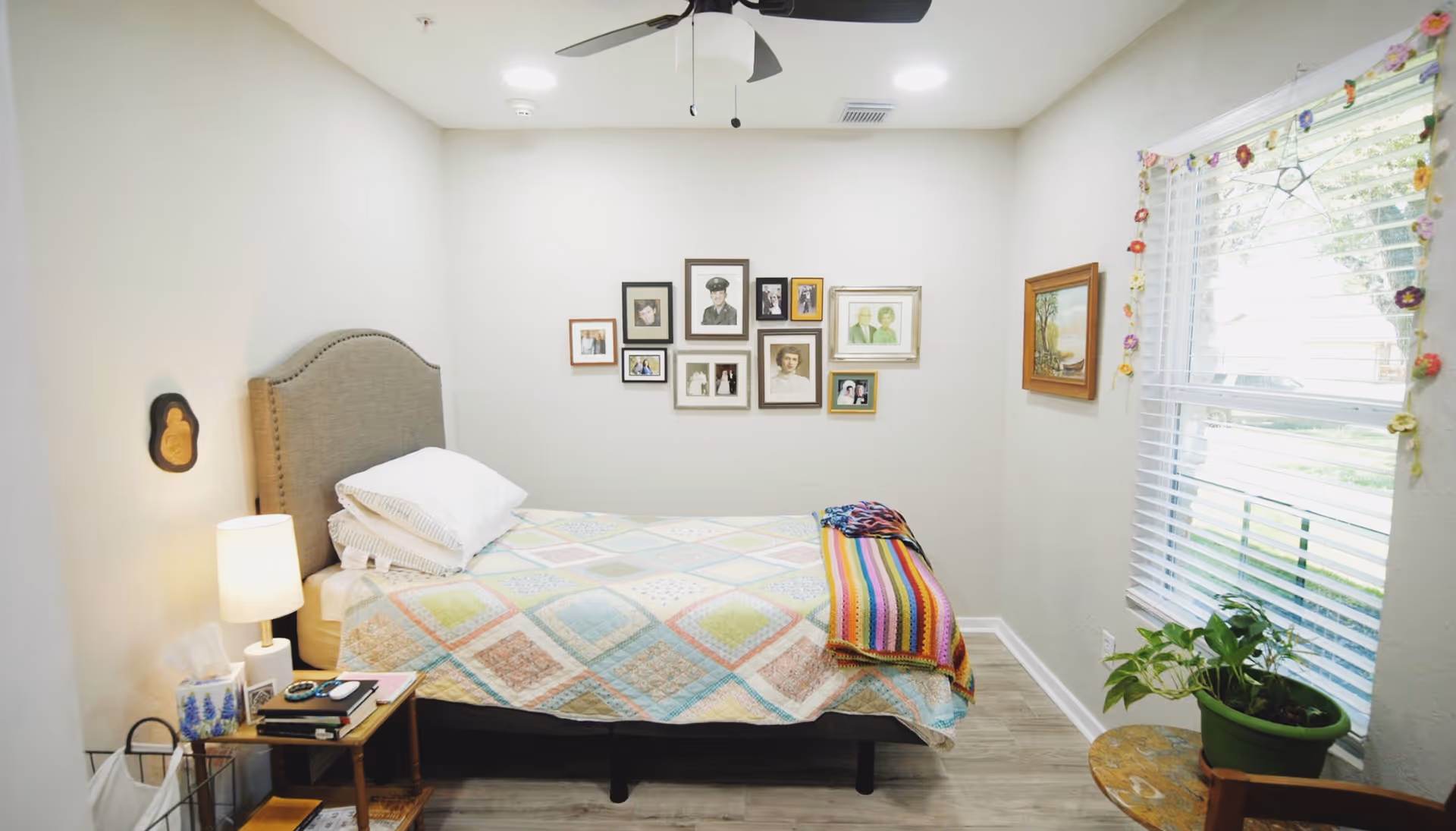 A cozy bedroom with a single bed covered in a colorful patchwork quilt and a striped blanket at the foot. There is a small wooden side table with a lamp, tissues, and books next to the bed. The walls are decorated with framed photographs and paintings. A window with white blinds and a floral garland lets in natural light. A green potted plant sits on a small round table near the window.
