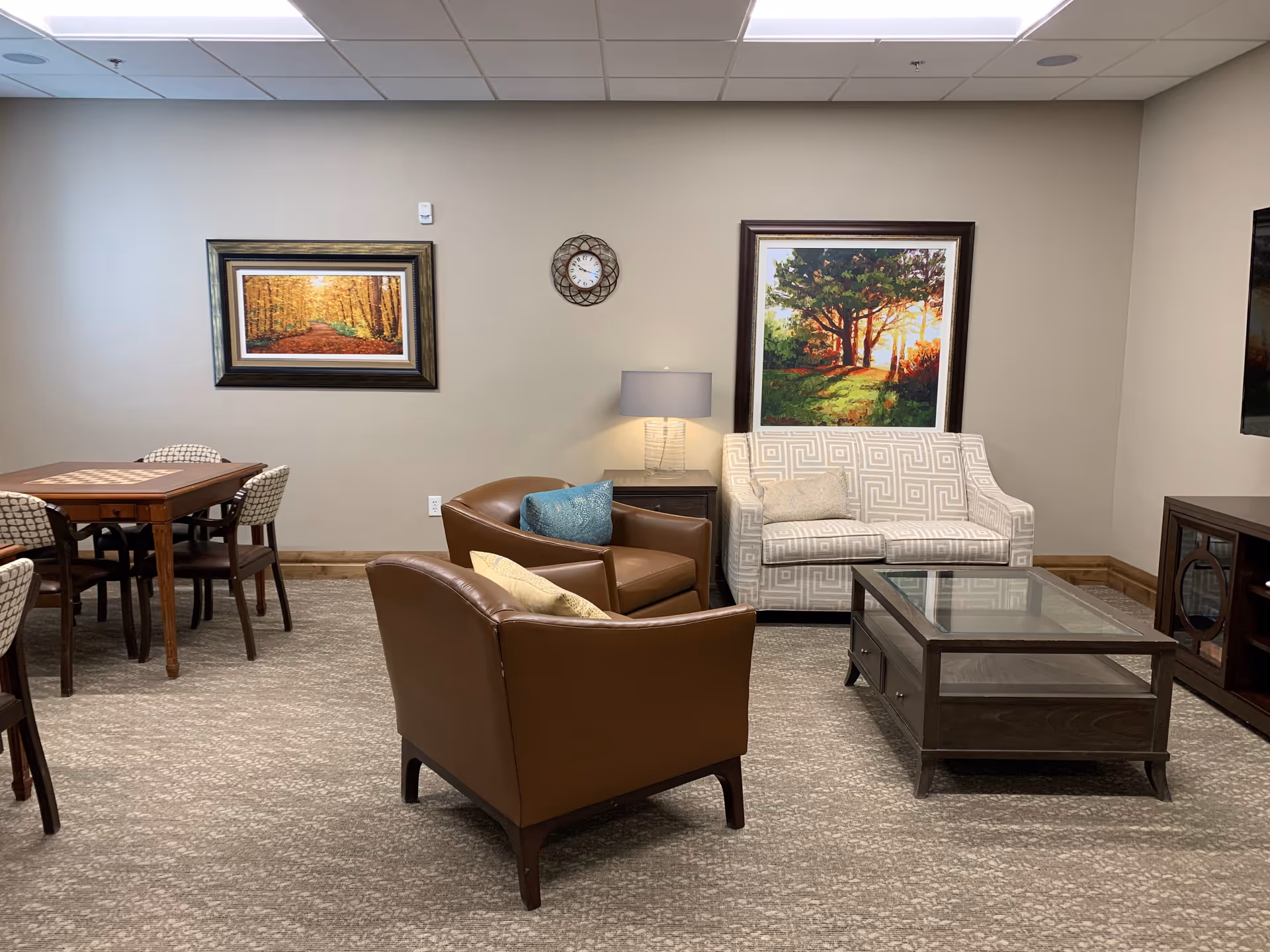 A cozy living room area with two brown leather armchairs, a white patterned loveseat, a glass coffee table, and a wooden side table with a lamp. There are two framed landscape paintings on the beige walls and a round wall clock. To the left, there is a wooden dining table with four chairs. The room has a carpeted floor and a drop ceiling with fluorescent lighting.