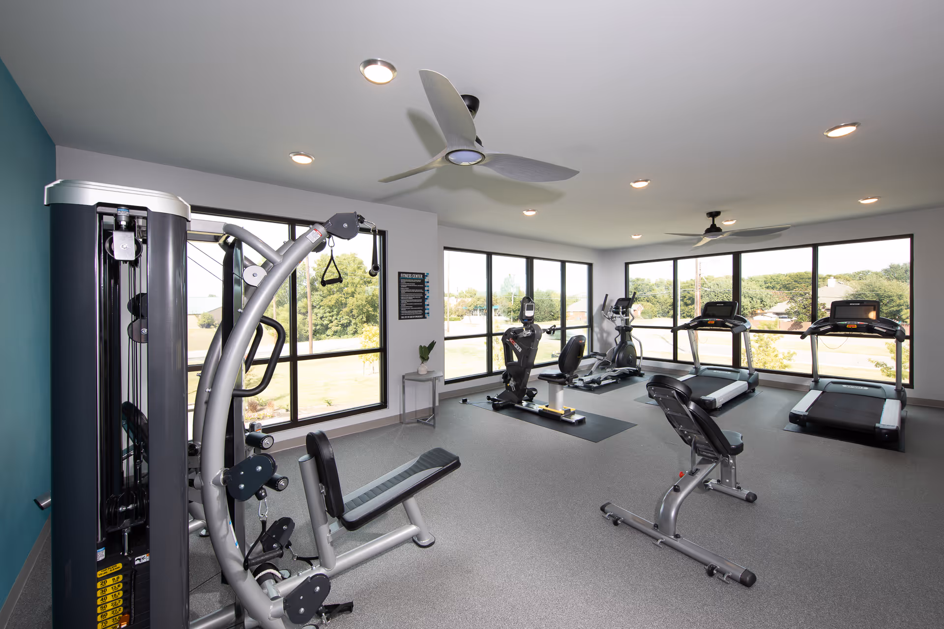 Well-lit exercise room with treadmills, stationary bikes, and weight machines in front of large windows.