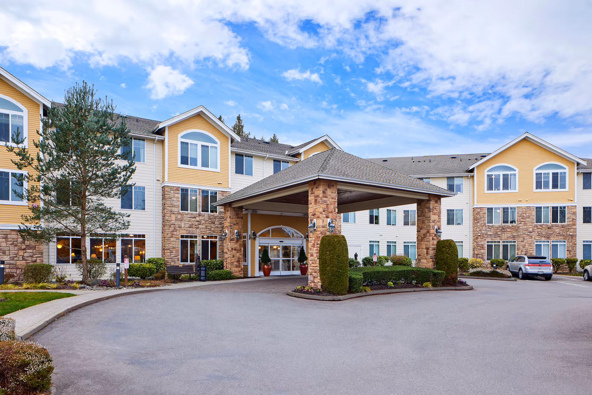 Exterior view of a senior living facility with a covered entrance supported by stone pillars. The building has three stories with a combination of stone and yellow siding, multiple windows, and a paved driveway with parked cars. The sky is partly cloudy.