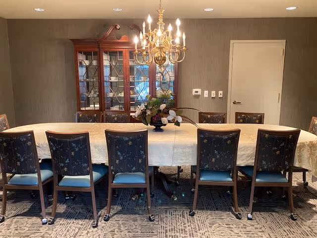 A formal dining room with a long rectangular table covered with a white tablecloth and surrounded by ten wooden chairs with blue cushioned seats and floral patterned backs. A brass chandelier with lit bulbs hangs above the table. Behind the table is a wooden china cabinet with glass doors displaying dishes. The walls are covered in textured wallpaper, and there is a closed white door on the right side.