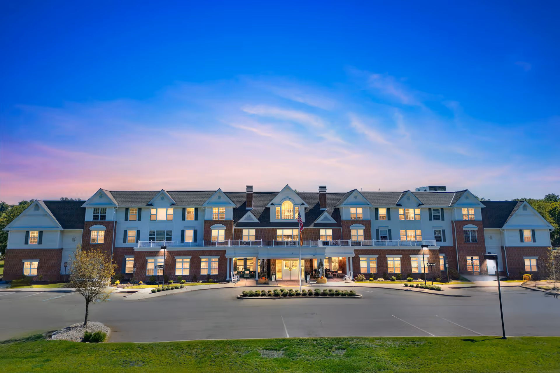 Exterior front view of a large three-story senior living facility building at dusk with lights on inside. The building features a combination of brick and white siding with multiple windows, a central entrance with a covered porch, and an American flag in front. The sky is clear with a gradient from blue to pink.