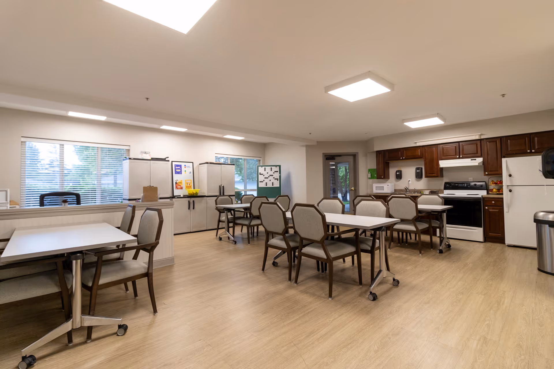 A spacious communal dining area with several tables and chairs arranged neatly. The room features large windows with blinds, light wood flooring, and a kitchen area with dark wood cabinets, a white refrigerator, stove, and microwave. The space is well-lit with ceiling lights and has a clean, organized appearance.