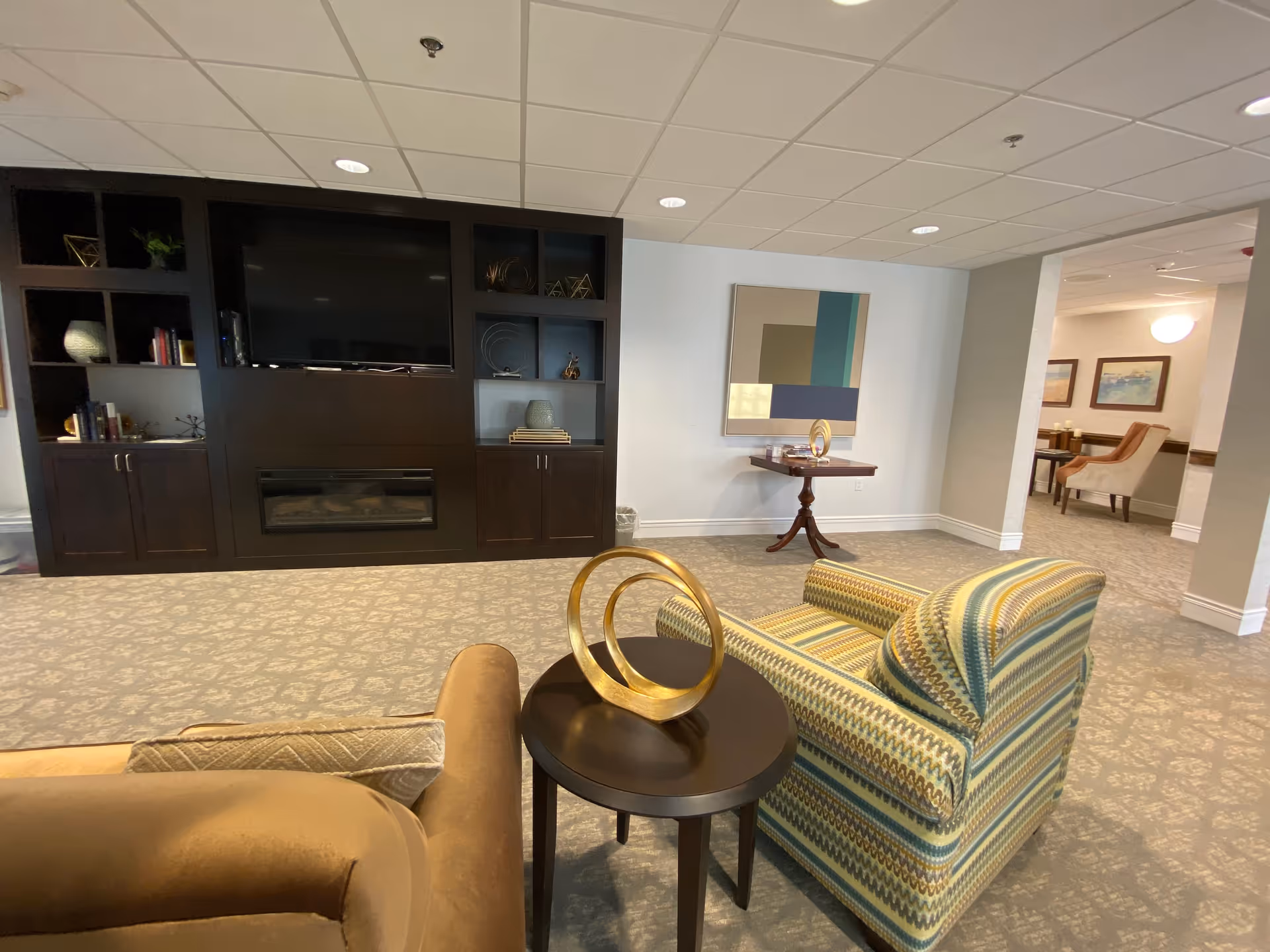 A cozy senior living common area featuring a patterned armchair and a brown sofa around a small round table with a gold decorative sculpture. In the background, there is a dark wood built-in entertainment center with a large flat-screen TV and a fireplace below it. A small wooden side table with another gold sculpture and a modern abstract painting hang on the wall. The carpet has a subtle floral pattern, and the ceiling has recessed lighting.