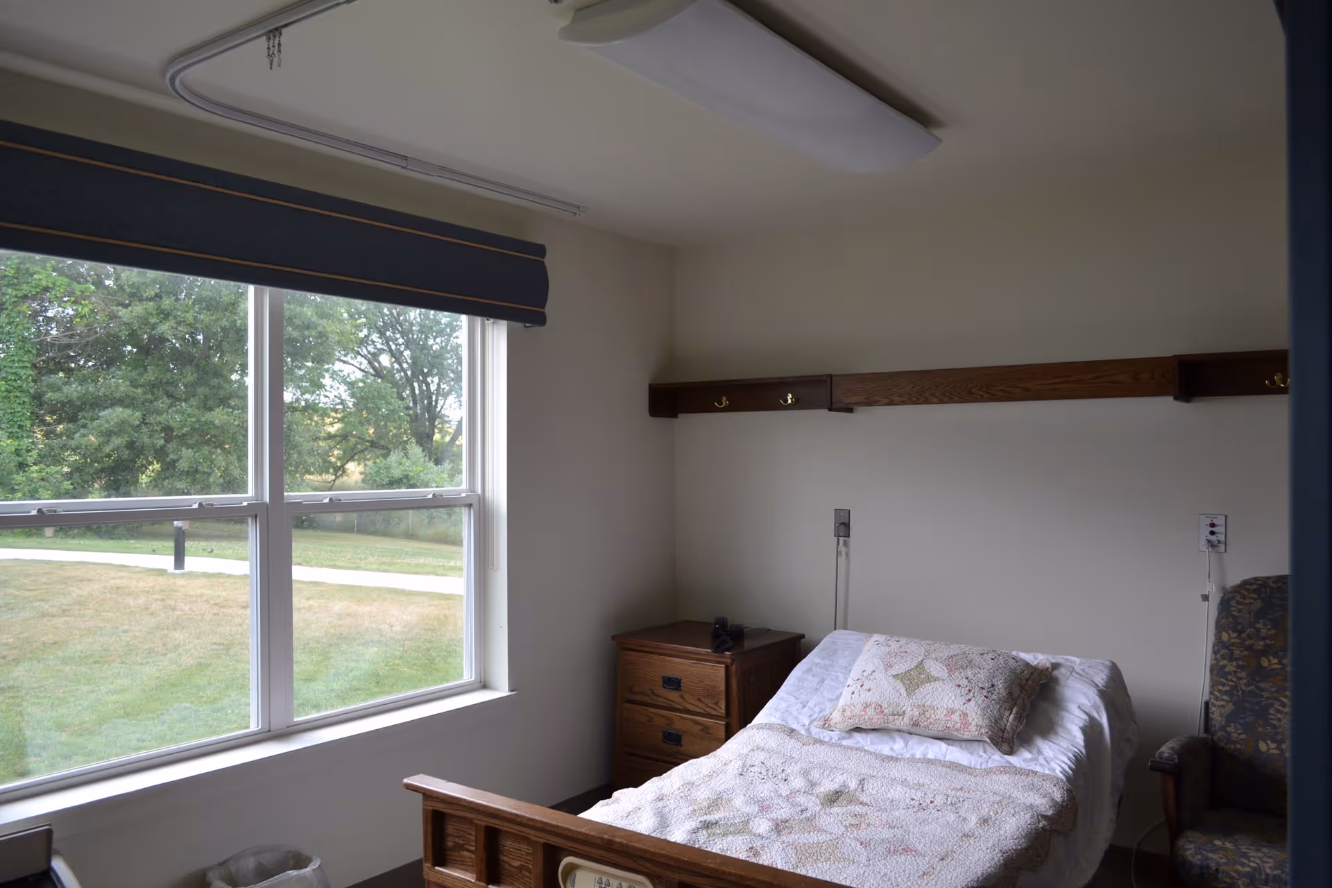 A small bedroom in a senior living facility with a single bed covered with a quilt and a pillow. There is a wooden nightstand next to the bed and a patterned armchair to the right. A large window with a dark blue valance lets in natural light and shows a grassy outdoor area with trees.
