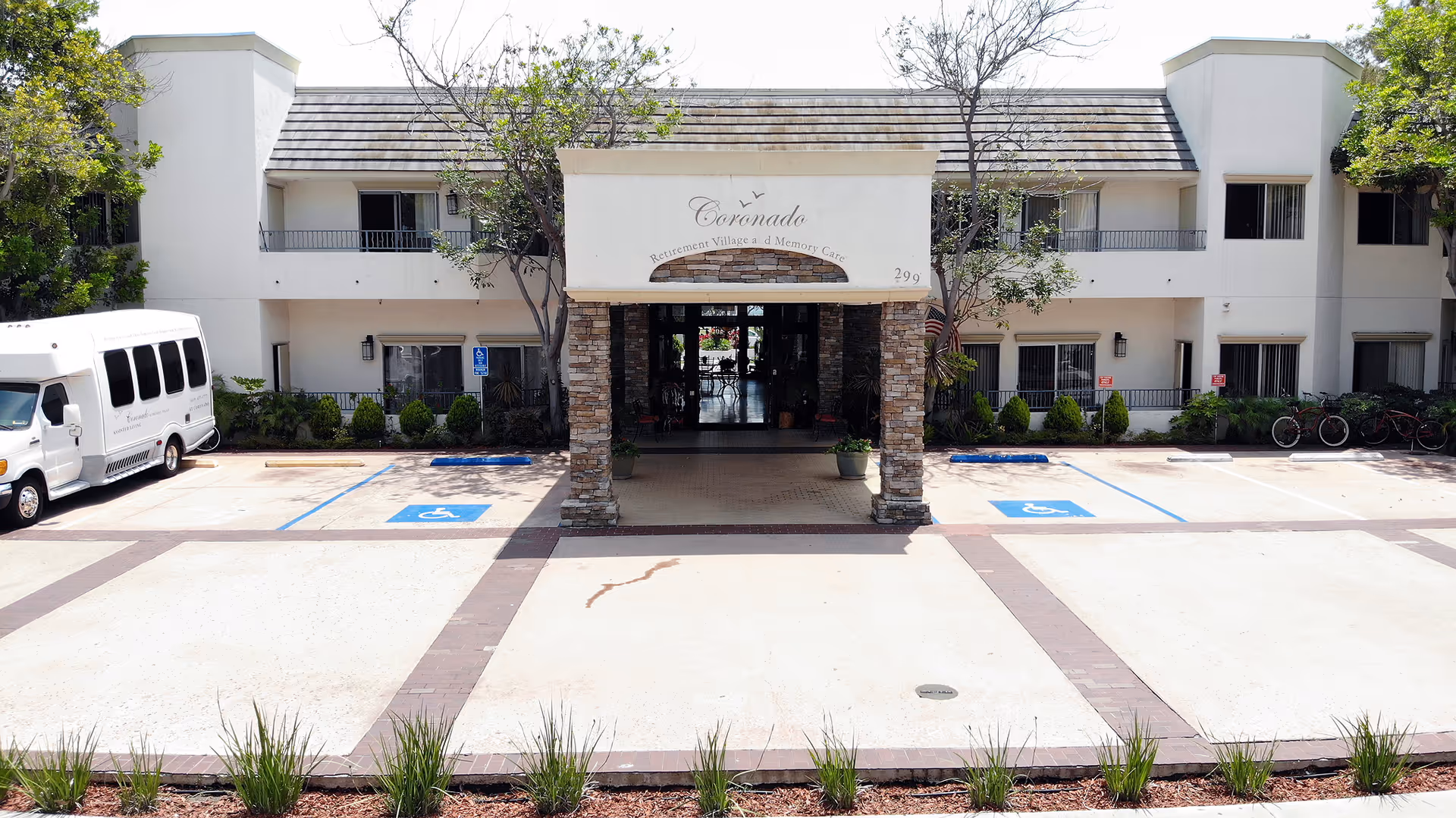 Front exterior view of Coronado Retirement Village building with a covered entrance supported by stone pillars, handicap parking spaces, a white shuttle bus parked on the left, and bicycles on the right side.