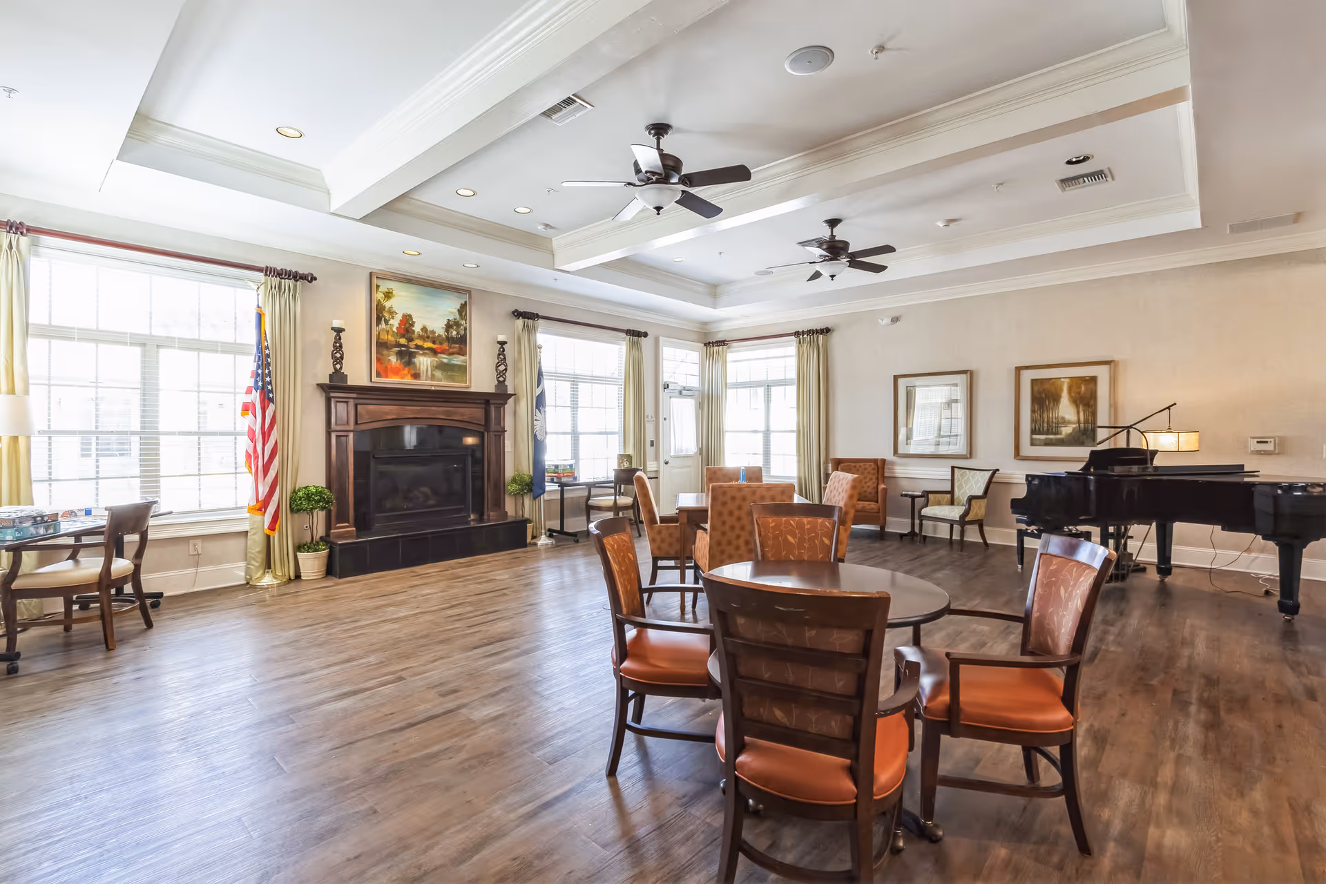 Bright senior living common room with round tables and chairs, a fireplace, large windows, and a grand piano.